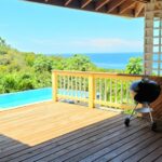 A wooden deck with a small black barbecue grill, an infinity pool, and a stunning ocean view in the background.