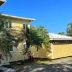 Yellow House Exterior: A bright yellow two-story house surrounded by tropical palm trees and plants under a clear blue sky.