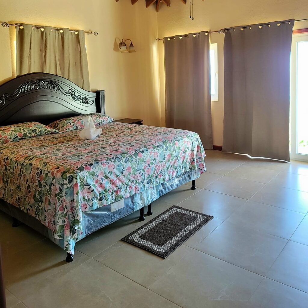 Spacious Bedroom with Floral Bedding: A large bedroom featuring a dark wooden headboard, floral bedding, beige curtains, and tiled flooring