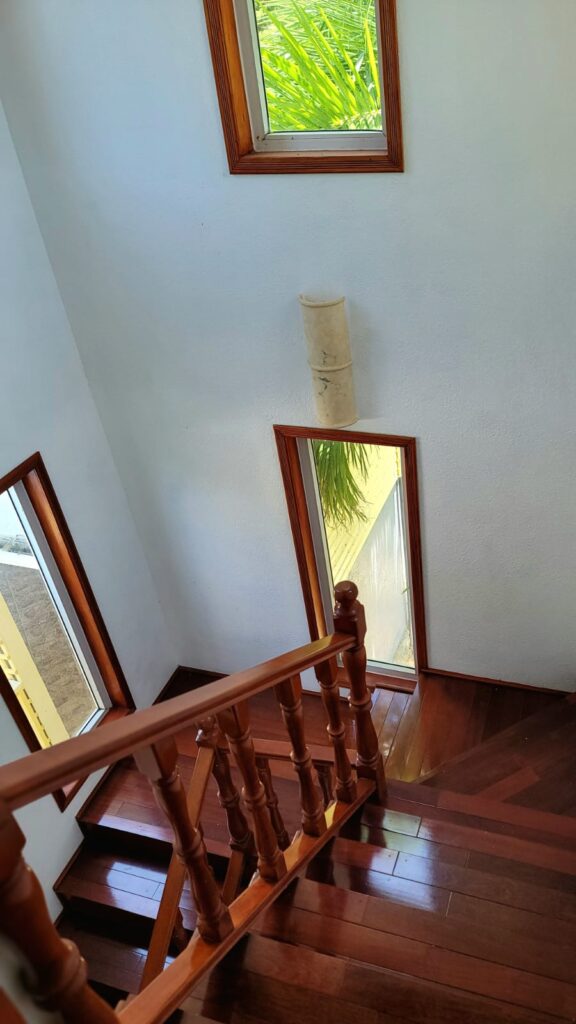 Interior Staircase View – A wooden staircase with a polished railing inside a well-lit house, featuring multiple windows with wooden trim