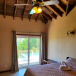 Bedroom with Balcony View: A cozy bedroom with a wooden ceiling, a ceiling fan, and a sliding glass door leading to a balcony overlooking a pool and greenery.