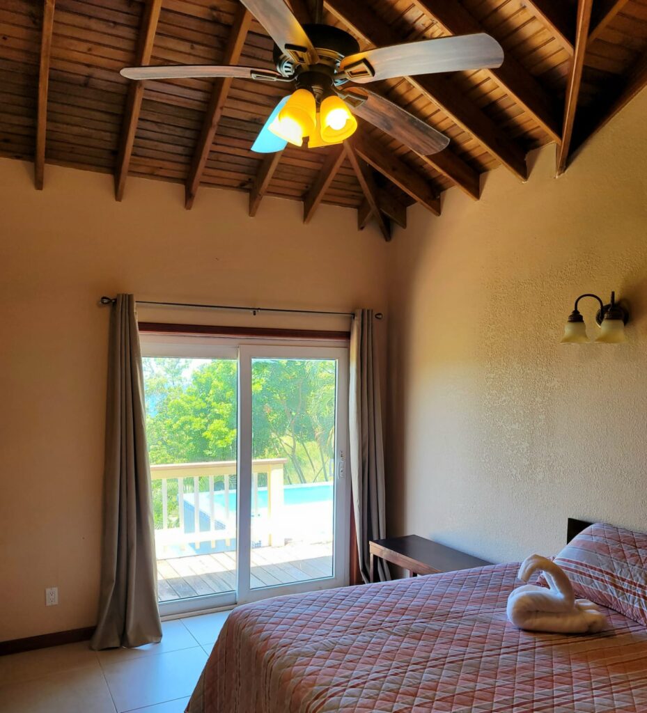 Bedroom with Balcony View: A cozy bedroom with a wooden ceiling, a ceiling fan, and a sliding glass door leading to a balcony overlooking a pool and greenery
