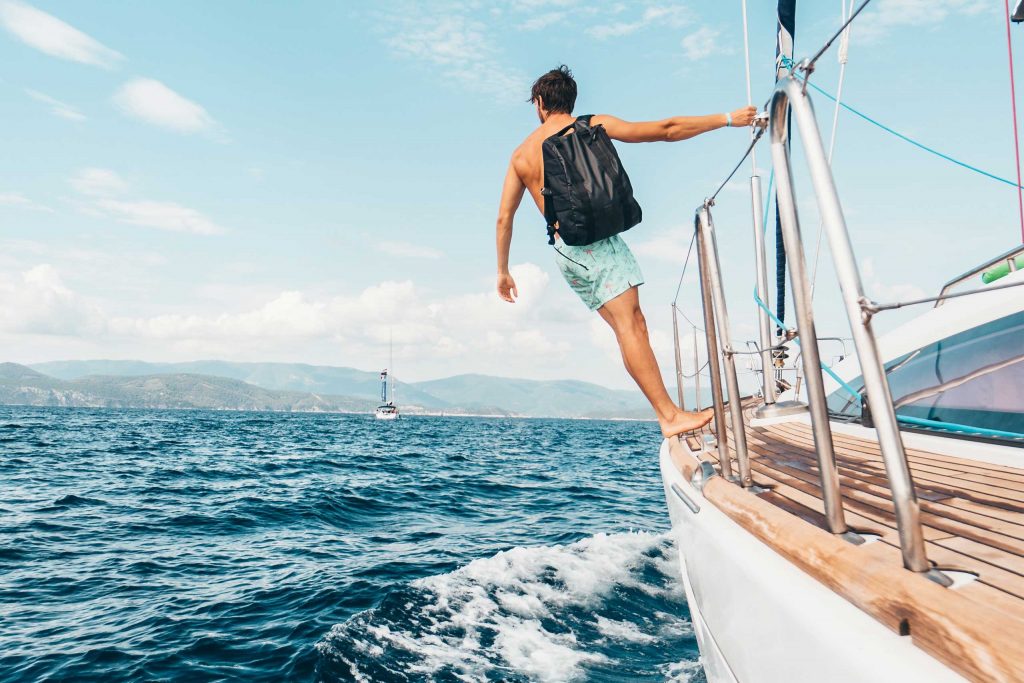 A man with a backpack standing on the edge of a sailboat, looking at the open sea.